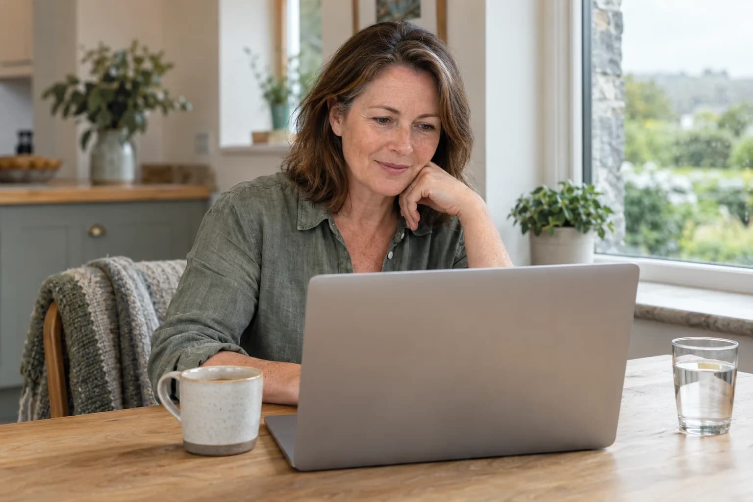 Mulher perto dos cinquenta anos numa consulta online à mesa da cozinha de uma casa irlandesa, com chá e água junto ao portátil e luz suave da janela.