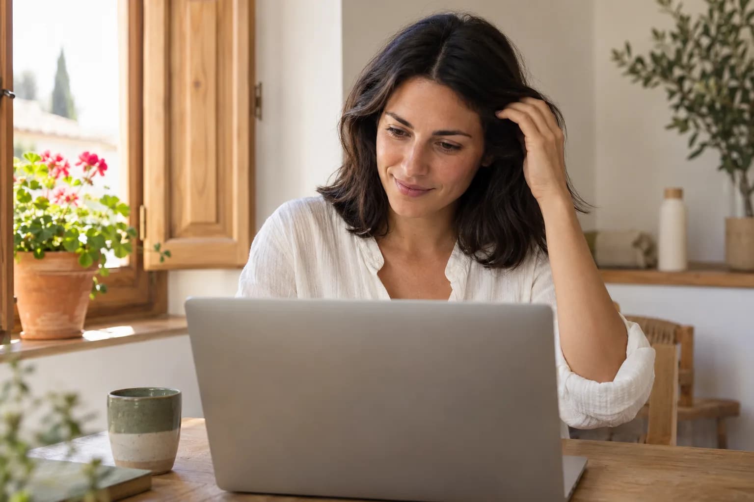 Žena si jemně sahá do vlasů a dívá se na notebook při online konzultaci, vedle slunného okna s dřevěnými okenicemi a muškáty.