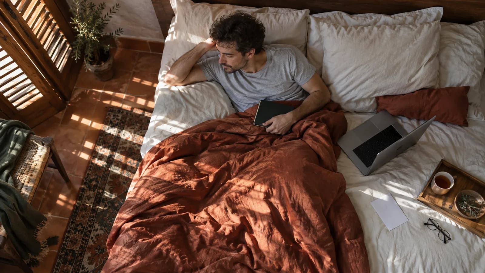 Man on sick leave resting in bed with a laptop and a herbal tea, seen from above.